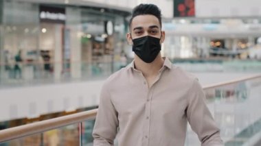 Millennial businessman rich man shopaholic wears medical face mask stands in shopping mall looking at camera happy showing money dollars holding package purchase bag with present buying sale discount