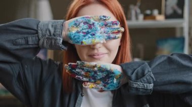 Female in art studio red-haired girl artist lady Caucasian 30s woman painter designer hides behind hands covers face with arms holding showing dirty in paint palms looking at camera smiling laugh