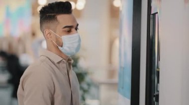 Millennial businessman man wears medical face mask uses bank terminal electronic machine for receiving and dispensing money ATM pays for purchase online order problems error currency exchange cash