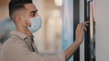 Millennial businessman man male client of cafe airport service in medical face mask uses bank terminal electronic machine for ATM for order buying online booking ticket pays for purchase with phone
