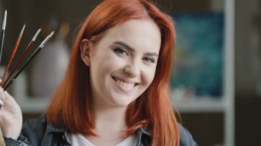 Female portrait in art studio red-haired 20s girl artist 30s woman painter creator designer looking at camera smiling holding drawing brushes in hand enjoying painting posing indoors with paintbrushes