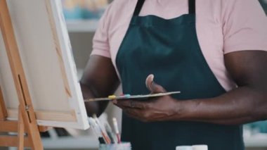 Close-up of male hands unrecognizable African man artist painter holding palette with bright oil acrylic paints colors drawing creates picture on canvas and easel using painting with brushes draw