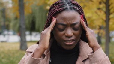 Close-up unhealthy frustrated woman holding head suffering from headache feels burning pain squeezing pressure suffers from chronic migraine unhappy girl stands outdoors feeling symptom of illness