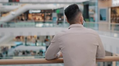 Millennial businessman man in formal shirt wears medical face protective mask stands in office business center in shopping mall looks at people public place during coronavirus epidemic pandemic covid