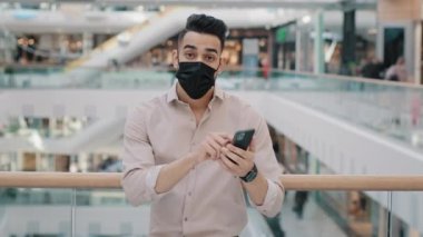 Millennial businessman man wears medical face mask stands in office center in shopping mall looking at camera displeased puzzled points to mobile phone broken smartphone problems with internet device