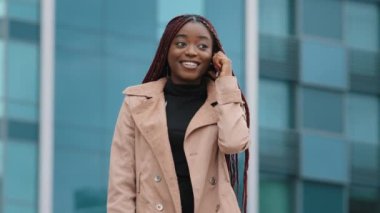 Young positive pretty african american woman standing against background of office building talking on phone answering mobile friendly call remotely chatting with friend using smartphone laughing