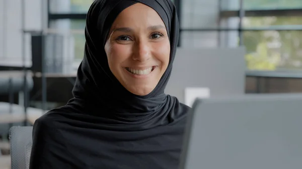 Close-up positive young woman office worker professional manager working on computer successful attractive arab businesswoman in hijab sitting at workplace looking at camera smiling enjoying workflow