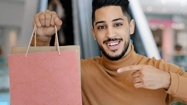 Happy young arab man buyer standing in mall smiling holding shopping bags in hand pointing forefinger at gift packages rejoicing at sale discounts showing ok sign approval gesture successful purchase