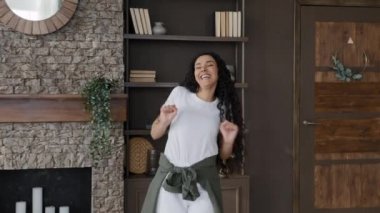 Young excited carefree woman dancing comic funny moves fooling around celebrating victory enjoying success happy joyful girl humorous moving jumping whirl falls on sofa having fun at home on weekend