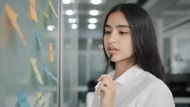 Arabian business lady managing daily tasks stand near board in office write reminder information instruction on paper sticky notes read stickers create to-do list analyze work organize corporate goals