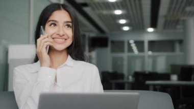 Busy female worker woman talk phone answer call arabic businesswoman do multitasking work on laptop chatting provide mobile support assistance for client in coworking office space make internet order