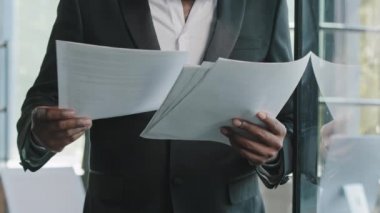 Unknown male businessman teacher boss auditor banker african man in black formal suit stands in office place check financial documents reading paperwork turn sheets banking notification loan insurance
