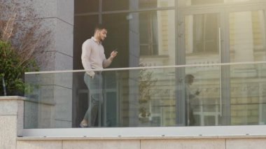 Walking businessman 30s pensive busy boss leader worker manager man guy walks on balcony terrace in company office building in hotel using smartphone working online remote with mobile phone chatting