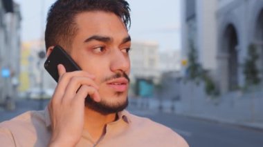 Close-up Hispanic Indian businessman bearded man talking mobile phone remote conversation distant talk answering call with smartphone calling ordering taxi standing in city street emotional dissuasion