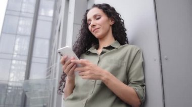 Confident 30s adult woman success businesswoman smiling texting in mobile app using phone online e-commerce service in internet chatting with smartphone outdoors leaning to wall of office building