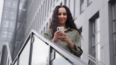 Smiling Caucasian Hispanic happy satisfied woman user lady standing un city outdoors on stairs chatting texting searching in net with mobile app phone browsing smartphone friendly communication online