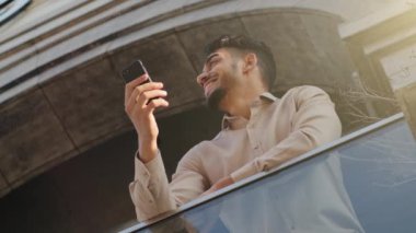 Bottom view cheerful smiling 30s bearded businessman male user man boss leader trader worker standing on balcony terrace at home office hotel using phone order taxi online app browsing with smartphone