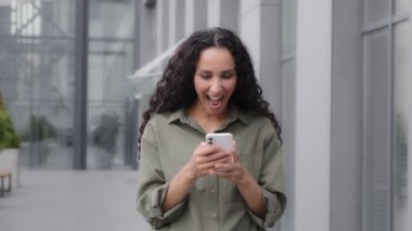 Adult woman 30s businesswoman entrepreneur female winning with good result news in cell phone standing outdoors success win opportunity internet bet achievement mobile app surprised lady celebrating