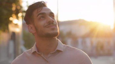 Close-up pensive inspired Indian Latin Arabic Hispanic bearded male boss worker man in formal shirt outdoors looking at sky thinking. Portrait businessman stands in city street in sunset sun dreaming