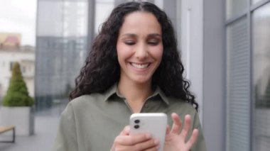 Latin Caucasian Hispanic smiling woman success businesswoman using cell phone smartphone app Wi-Fi device outdoors walking going in city near office browsing chatting texting with colleagues online