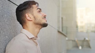 Close up side view pensive dreaming inspired bearded Hispanic Indian adult 30s businessman relaxed outdoors man resting take break standing leaning wall in city looking up in sky dream contemplating