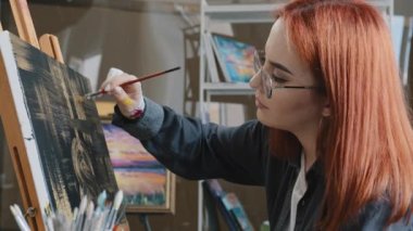 Inspired busy concentrated Caucasian red-haired woman in eyeglasses girl painter wear glasses painting drawing on canvas with yellow gold paints practicing hobby art education skill draw with brush