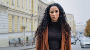 Ciddi, yalnız, kendine güvenen, uzun kıvırcık saçlı, kendine güvenen, genç, esmer bir kız. Yüzünde kızgın bir ifade olan iş kadını şehirde dikilip kameraya bakıyor.