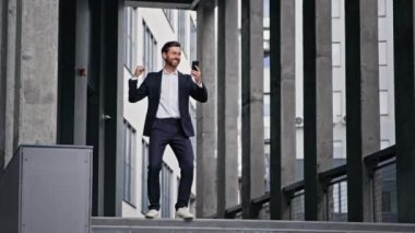 Young stylish businessman stands against backdrop of office building reads good news on telephone celebrates victory rejoices in success business promotion achievement excellent result receives reward