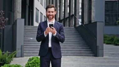 Young successful businessman standing outdoors in business center holds phone ordering taxi using mobile application checking email browsing social network chatting to client on smartphone writing sms