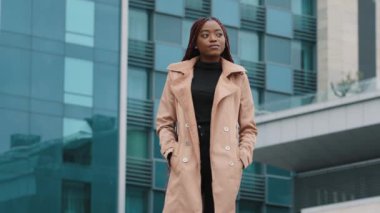 Confident young african american businesswoman standing in front of office building waiting for meeting attractive fashionable girl model with stylish hairstyle posing outdoors looking around pending