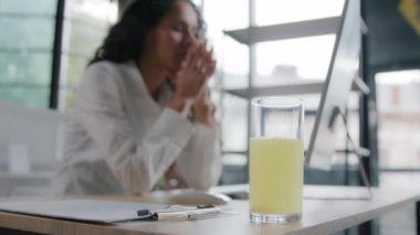 Close-up focus on glass with remedy aqueous liquid with painkiller unhealthy tired woman worker sitting at workplace feels headache suffering from chronic migraine concept treatment with medical drug
