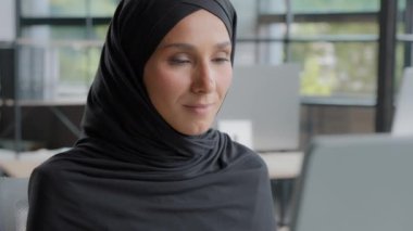 Close-up positive young woman office worker professional manager working on computer successful attractive arab businesswoman in hijab sitting at workplace looking at camera smiling enjoying workflow