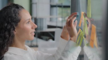 Side view smiling happy successful businesswoman taking notes on stickers writing reminders on color sticky label joyful female project manager developing startup strategy creates plan on glass board