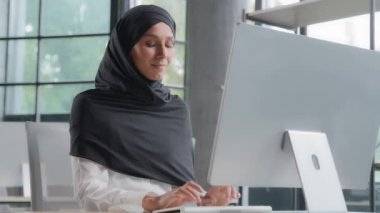 Young arab businesswoman in hijab working on computer smiling enjoying office work successful woman professional manager company employee develops project online searching for information on Internet
