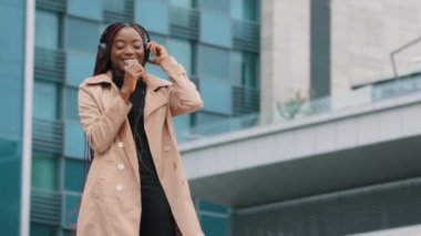 Happy carefree stylish young african american woman listening to music on smartphone with headphones outdoors joyful girl dances sings song uses phone as microphone active moving favorite audio track
