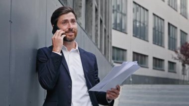 Young confident executive businessman stands outdoors talking on mobile phone holding paper documents consulting client using smartphone answers call discussing business matters on telephone on street