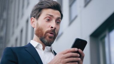 Close-up young surprised man looks at phone screen receiving notification reading good news rejoicing in success satisfied businessman gets excellent result from sales profitable offer salary increase