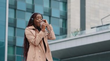 Joyful energetic young woman dancing outdoors listening to music with headphones singing using smartphone as microphone funny girl performs active dance movement listens to favorite song in earphones