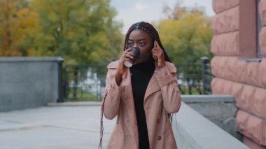 Serious young woman walking outdoors drinking hot coffee from paper cup talking on telephone answering business call stylish businesswoman communicates remotely with client using smartphone on street