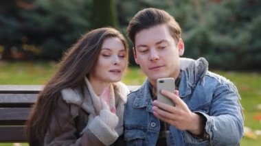 Caucasian couple young family partners sit on bench in autumn park look at phone camera wave hand hello communicate distant with friends smiling talk online chat joint internet meeting use cellphone