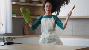 Playful comical woman arabian girl suddenly appears jump in the kitchen surprise effect funny startle wears oven mitt hold wooden spoon play game with children child trick has fun domestic amusement