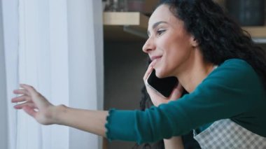Smiling young arabian woman chatting with friend by cellphone has pleasure phone conversation gossip stand in house look at window greet friend from outside quarantine self-isolation due covid concept