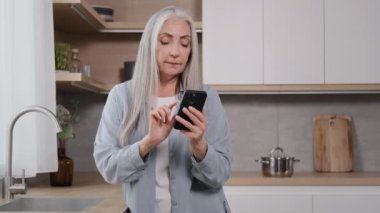 Excited happy elderly woman amazed female grey-haired granny pensioner housewife winner stand at home kitchen read good news on mobile phone rejoice online bet victory win discounts in internet store