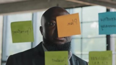 African american analyst serious businessman project manager check schedule read information on reminders stickers look at glassy wall with colorful sticky notes managing goals daily corporate tasks