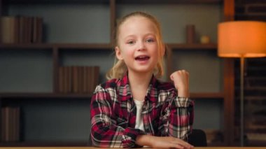 Little funny happy blonde school kid toddler child girl five years old winner in red shirt clenching fist doing gesture say yes celebrating success winning goal achievement lottery luck at home room