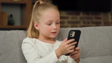 Beautiful pretty small girl pupil caucasian kid child daughter blogger sit on sofa at home use smartphone play mobile games chatting with classmates in social networks watching funny cartoons at home