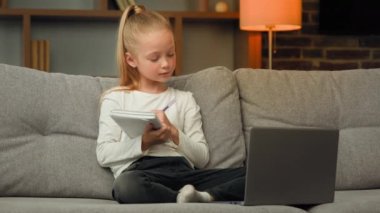 Blonde little smart schoolgirl pupil listen educational lesson on laptop at home online class meeting in video chat during pandemic write homework tasks at copybook raise hand ready to answer question