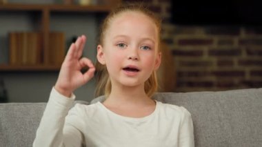 Portrait of caucasian happy child girl smiling kid sitting on sofa at room looking at camera showing ok gesture everything fine like sign positive good emotion. Little cute charming teen kid recommend