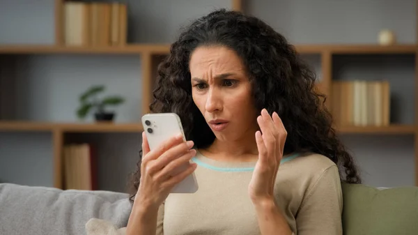 Hispanic sad displeased woman has problem with online mobile app on cell phone unhappy with blocked account in social networks looking at cellphone screen scam unsuccessful bad news sitting at home