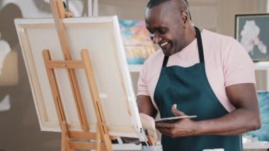Side view happy smiling cheerful creative talented inspired old African American senior man artist male painter in apron drawing painting picture creative idea on canvas in art studio school paints